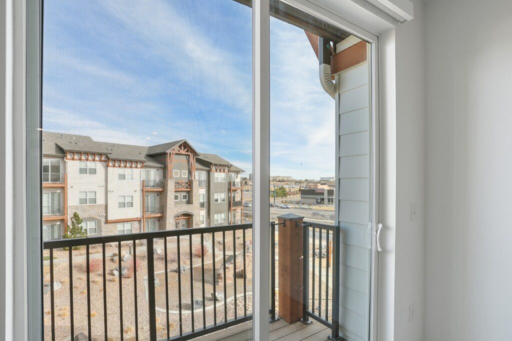 View from inside an apartment, looking out through a glass sliding door onto a small balcony with a black railing. An adjacent apartment building and a landscaped area are visible under a blue sky.