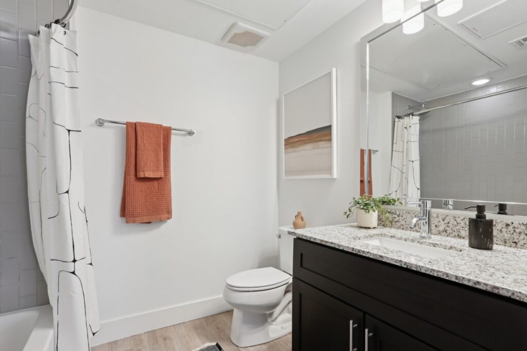 Modern bathroom with white walls, a shower with a patterned curtain, a toilet, granite countertop vanity with dark cabinets, a plant, orange towels, abstract wall art, and a large mirror above the sink.