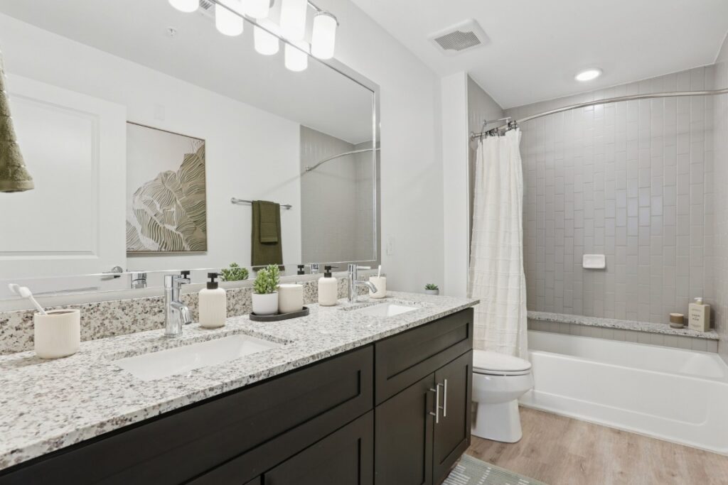Modern bathroom with double sink vanity, granite countertop, large mirror, potted plants, bathtub with tiled wall, white shower curtain, toilet, and wall art. Bright lighting and neutral color palette throughout.