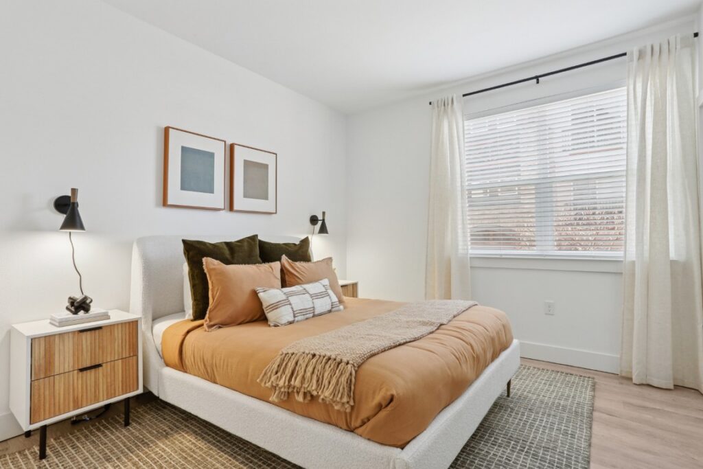A modern bedroom with a beige bed, tan bedding, two pillows, a throw blanket, two wall-mounted lamps, two framed artworks, a nightstand, and a window with white curtains letting in natural light.