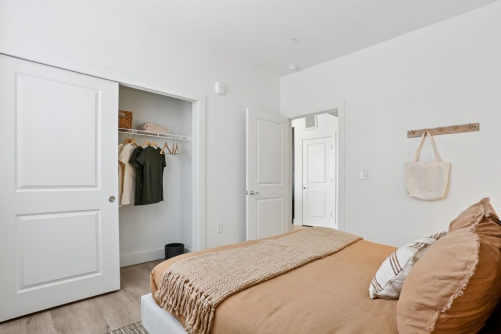 A cozy bedroom with a neatly made tan bed, pillows, and a beige throw. An open closet shows hanging clothes, baskets, and shelves. A tote bag hangs on the wall. White walls and doors create a bright, clean look.