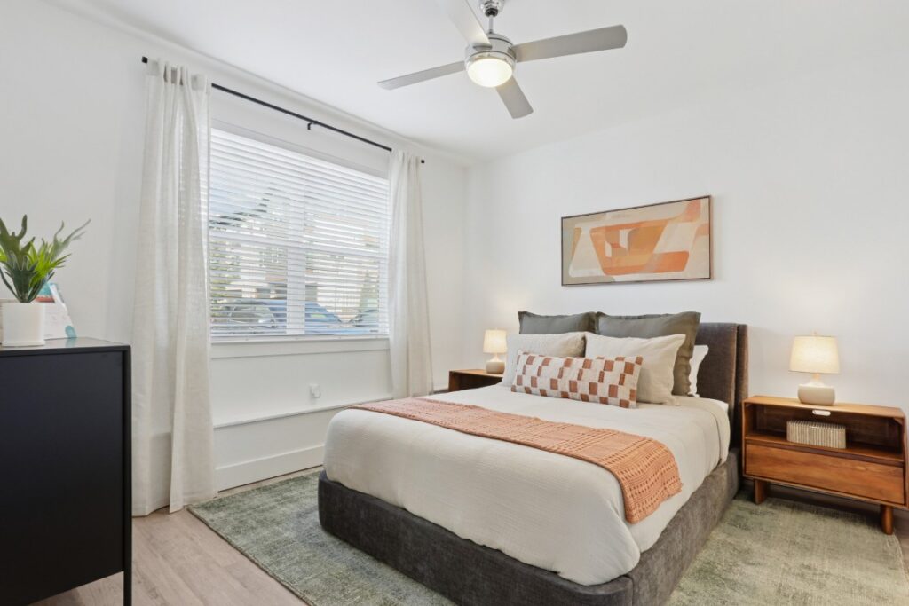 A modern bedroom with a bed, two nightstands with lamps, a dresser, and a large window with white curtains. Abstract art hangs above the bed, and a ceiling fan is overhead.