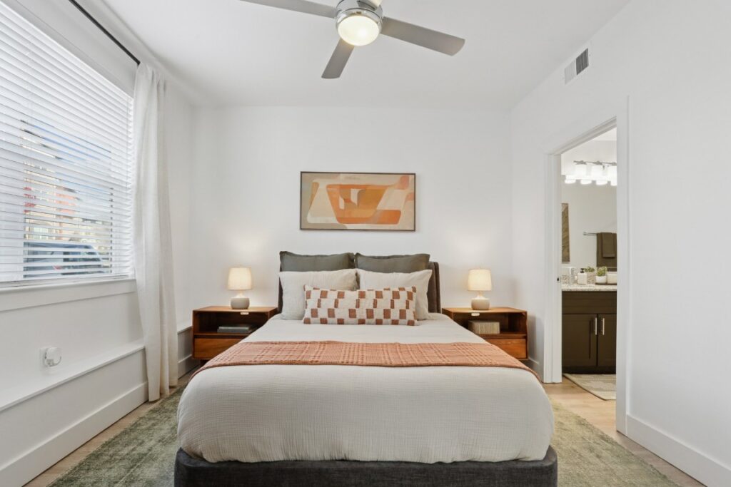 A modern bedroom with a neatly made bed, two bedside tables with lamps, a ceiling fan, large window with blinds, abstract artwork above the bed, and an open door leading to a bathroom.