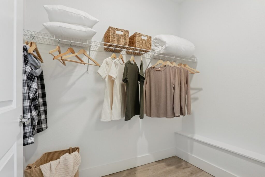 A tidy walk-in closet with clothes on wooden hangers, wicker baskets, and folded bedding on a wire shelf. Laundry hampers and a bench sit on a wood floor against white walls.