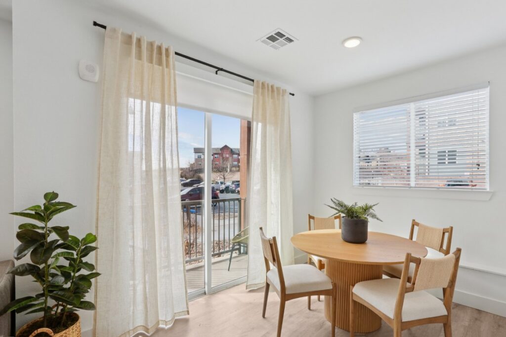 A small dining area with a round wooden table, four chairs, a potted plant centerpiece, sheer curtains on a sliding glass door, a window with blinds, and a green plant in the corner.