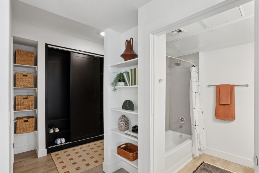Open bathroom with a white tub and orange towel, adjacent to hallway shelving with decor, baskets, and a black closet with shoes. Light wood floor throughout.