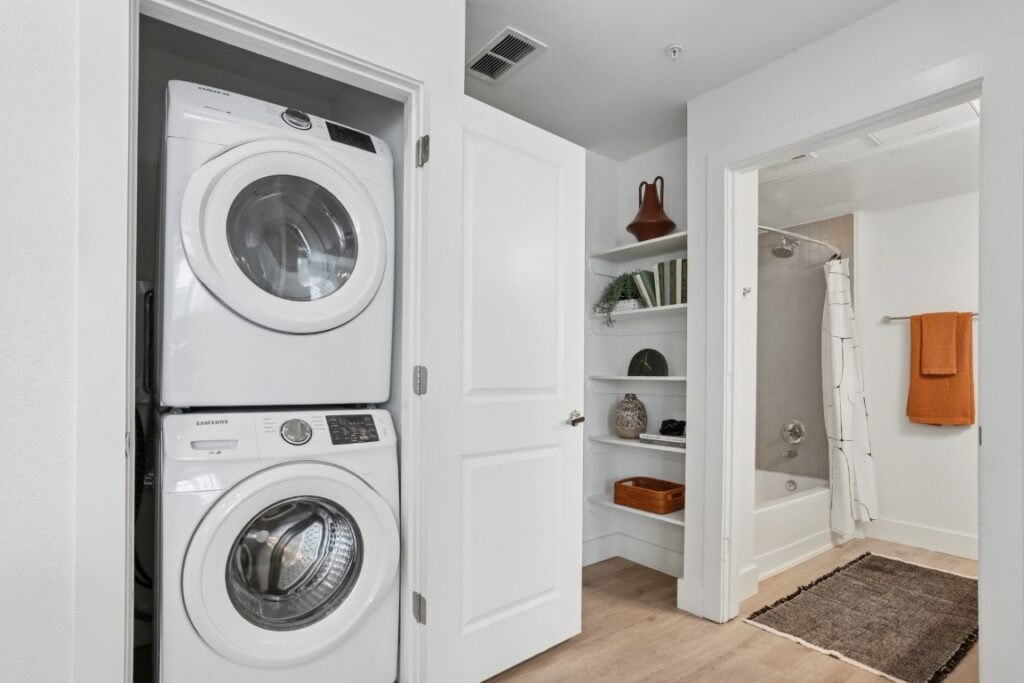 A laundry area with stacked washer and dryer next to an open door leading to a bathroom with shelves, a bathtub, and orange towels hanging on the wall.