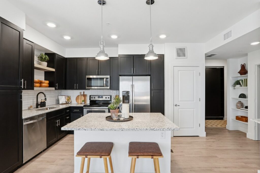 Modern kitchen with dark cabinets, stainless steel appliances, a white granite island with two wooden stools, light wood flooring, and open shelves with decor. Bright lighting and a clean, minimalist design.