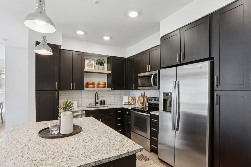 Modern kitchen with dark cabinets, stainless steel appliances, a granite island countertop, two pendant lights, and decorative items on open shelves against a white tile backsplash.