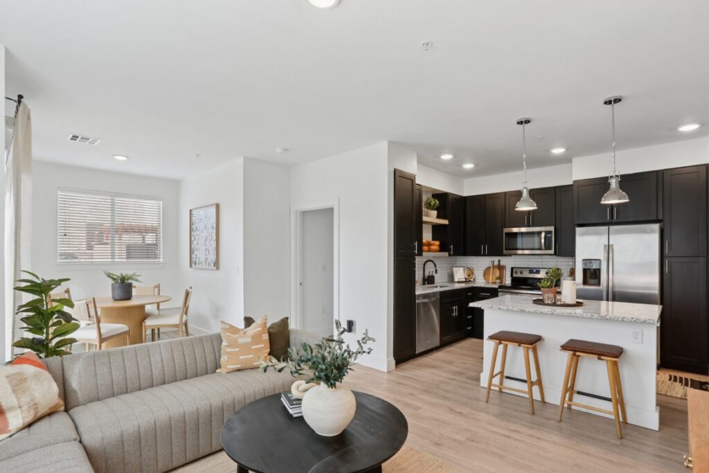 Modern open-concept living space with a gray sofa, round coffee table, dining area by a window, and a kitchen featuring dark cabinets, stainless steel appliances, and a breakfast bar with two stools.