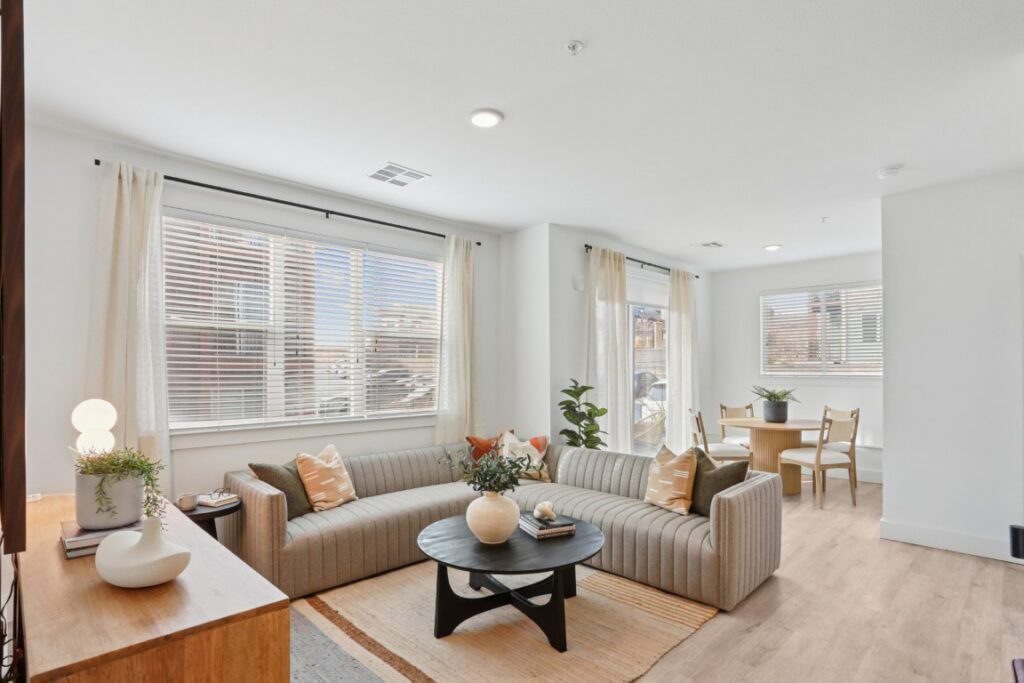 Bright, modern living room with a striped sectional sofa, round coffee table, plants, and decor. Large windows let in natural light. A dining table with chairs is visible in the background.