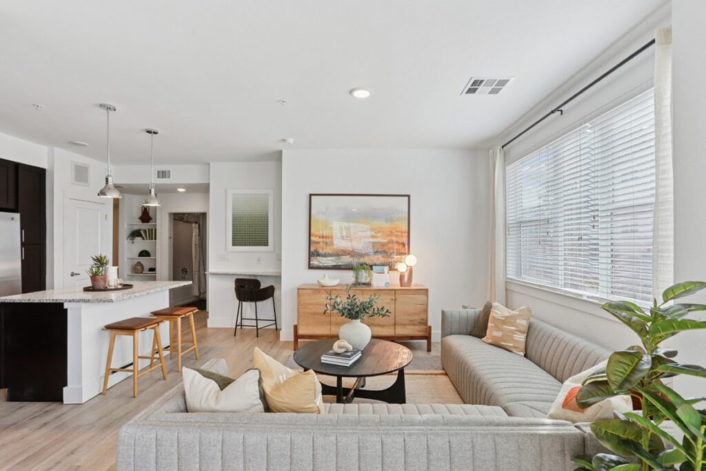 Bright modern living room with a gray sectional sofa, round coffee table, kitchen island with stools, and large window with white blinds. Art and lamps decorate a wooden sideboard against a white wall.