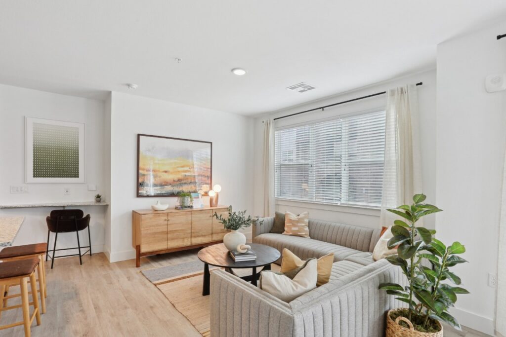 A bright living room with a gray sectional sofa, round coffee table, potted plant, sideboard with art and lamps, barstools at a counter, and large windows with white curtains.