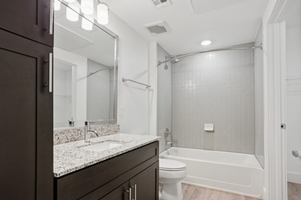 Modern bathroom with a large mirror, dark wood vanity with granite countertop, single sink, toilet, and bathtub with gray tile shower surround. Bright overhead and vanity lighting.