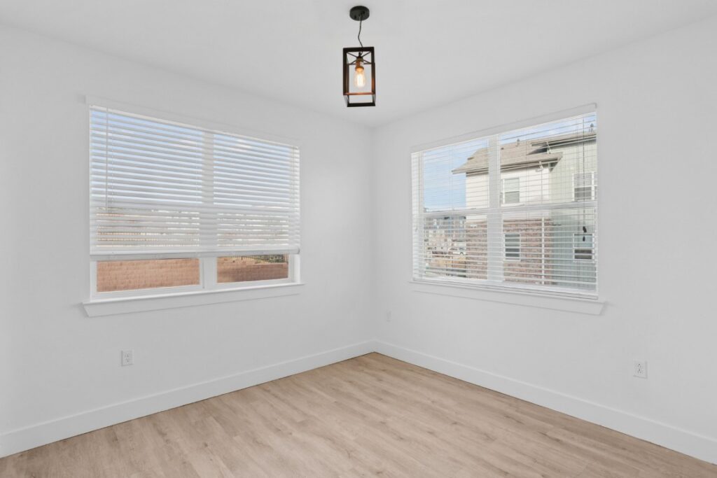 Bright, empty room with white walls, two large windows with blinds, light wood flooring, and a modern black ceiling light fixture. House exterior visible through the windows.