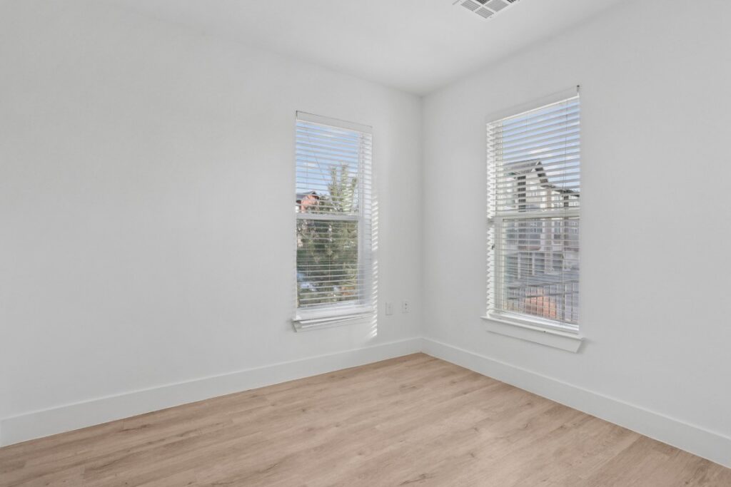 Bright, empty room with light wood flooring, white walls, and two tall windows with white blinds letting in sunlight. Some buildings and trees are visible outside.