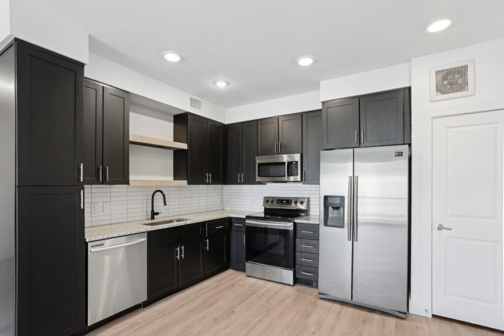 Modern kitchen with black cabinets, stainless steel appliances, light wood floor, white tile backsplash, open shelves, and recessed lighting. A white door and framed art are visible on the right side.