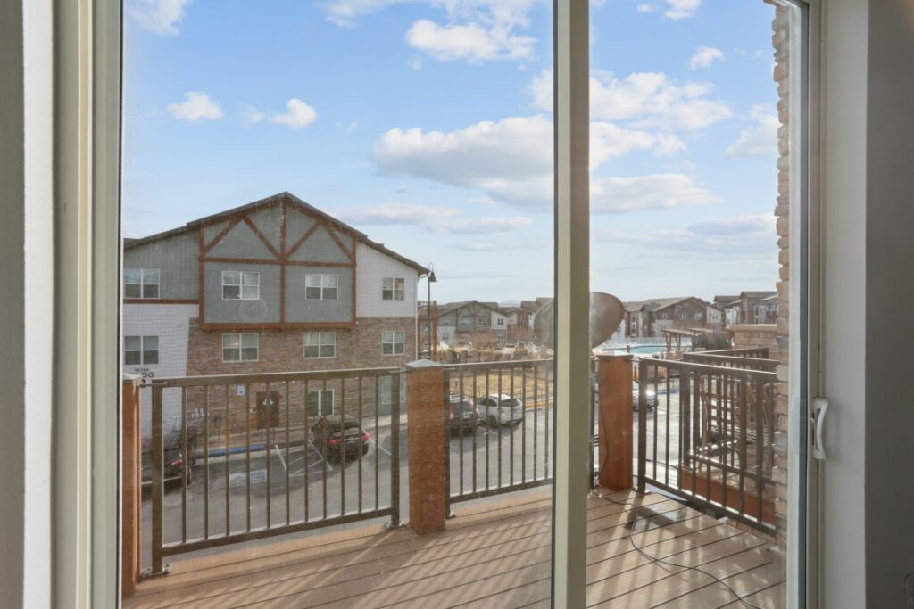 View through a glass door onto a balcony with a metal railing, overlooking an apartment complex under a blue sky with scattered clouds.