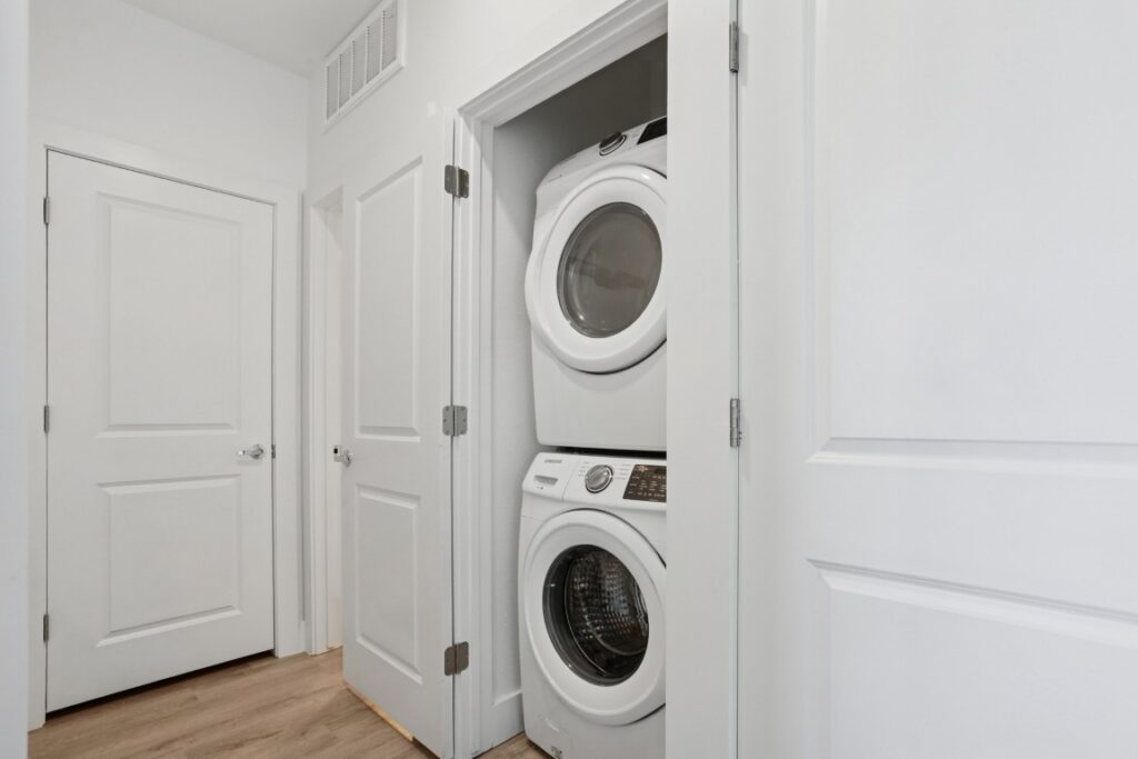 A small laundry closet with a stacked white washer and dryer, surrounded by white doors and walls, and light wood flooring.