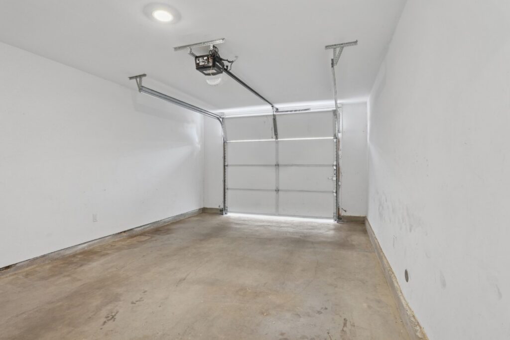 Empty, clean garage with white walls, concrete floor, ceiling light, and an automatic garage door partially open. Some scuff marks are visible on the lower part of the right wall.