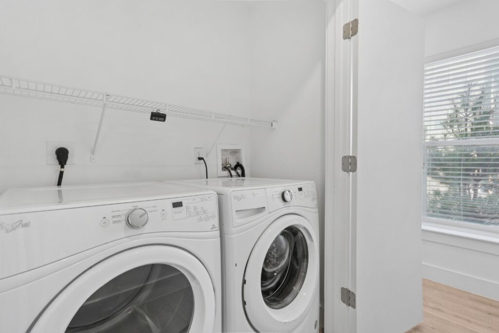 A laundry area with a white Whirlpool washing machine and dryer, white walls, a wire shelf above them, and a window with blinds letting in natural light.